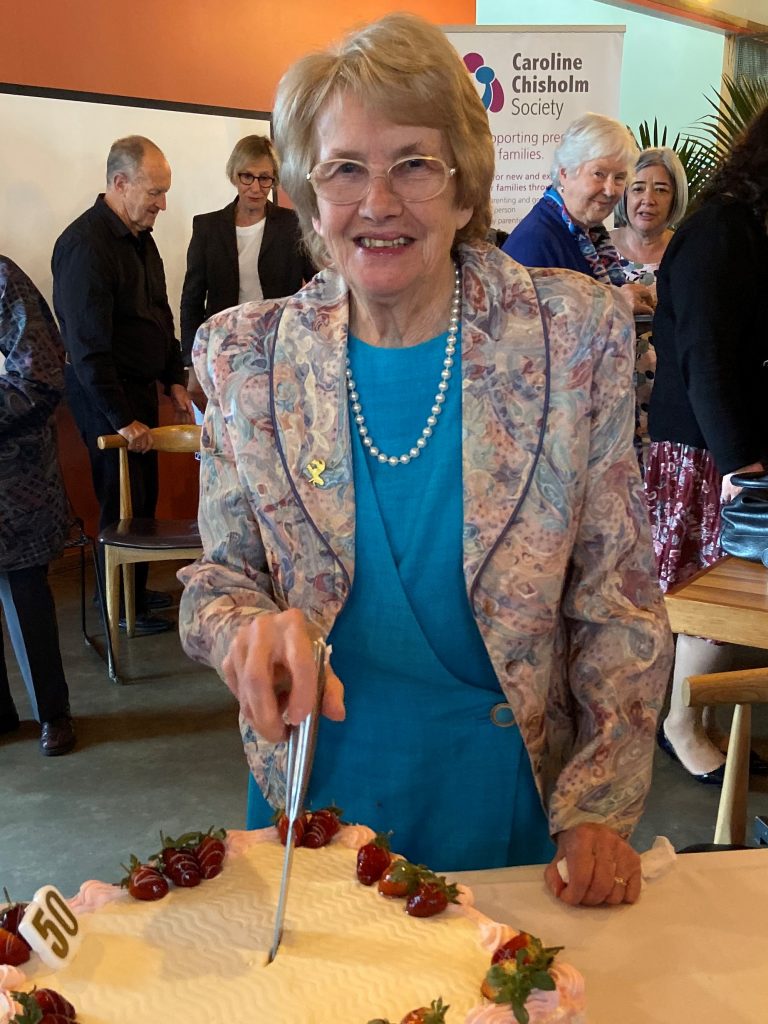 Dr Philomena Joshua wears glasses, a blue dress and floral blazer as she stands cutting a cake and smiling for a photo