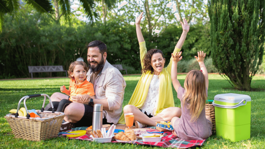 A family of four sit on a picnic rug in the park, with a picnic basket, food and tableware arranged between them. On the left side of the picnic rug, the a young boy sits on the fathers lap, while on the right a young girl and her mother sit facing each other, grinning with their arms raised in the air.
